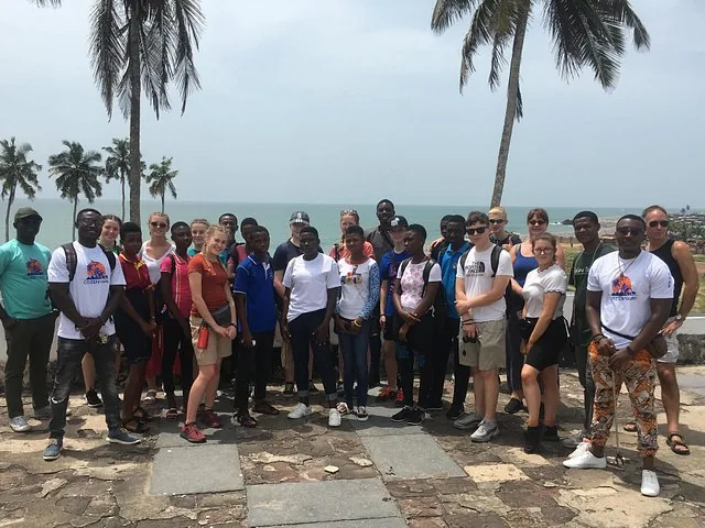 Large group of tourists posing for a photo overlooking the ocean near palm trees.